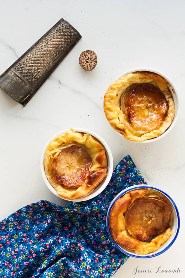 Ramekins of apple clafoutis baked until golden brown with a blue floral linen and half a nutmeg and a grater