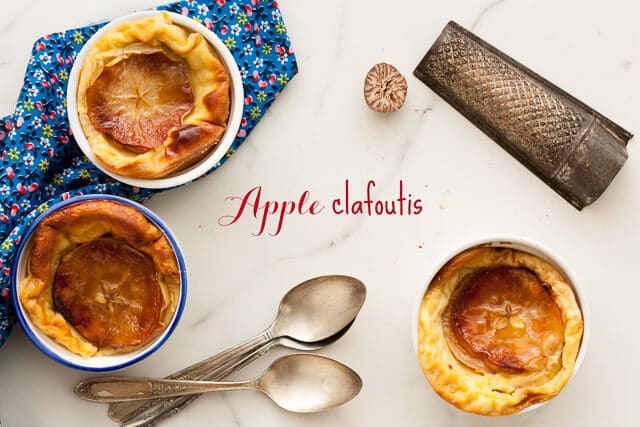 Baked apple clafoutis in individual ramekins with a nutmeg and grater, blue floral linen and spoons to serve on the side.