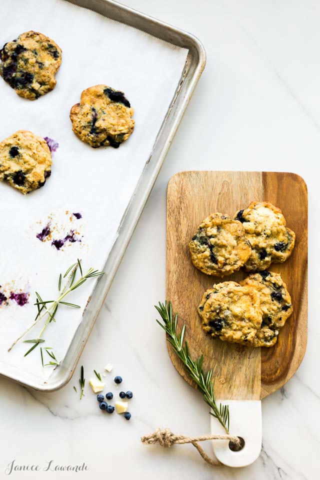 Oat blueberry white chocolate cookies with rosemary freshly baked on a parchment-lined sheet pan, transferred to a wood cutting board with marble handle and rope. Sprigs of rosemary, little blueberries and white chocolate on the side