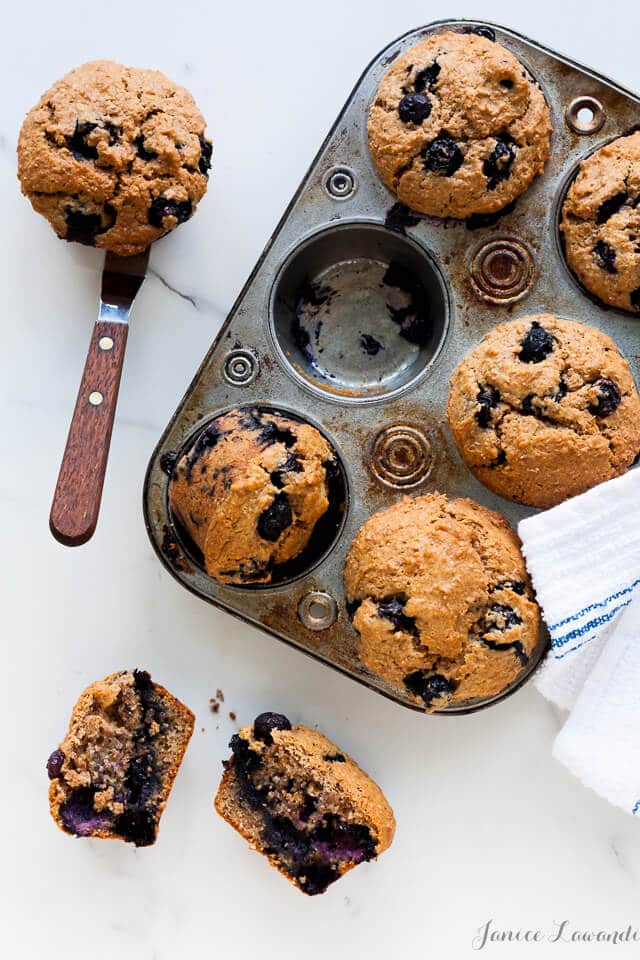 A muffin pan of blueberry bran muffins with 1 cup empty. One muffin on an offset spatula and another muffin broken open to reveal the interior