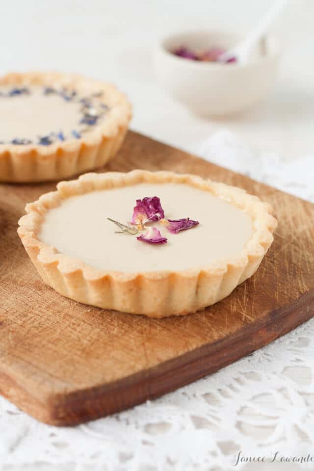 Little Earl Grey panna cotta tarts topped with dried rose petals and dried cornflower petals, served on a wooden cutting board placed on a white lace doily 