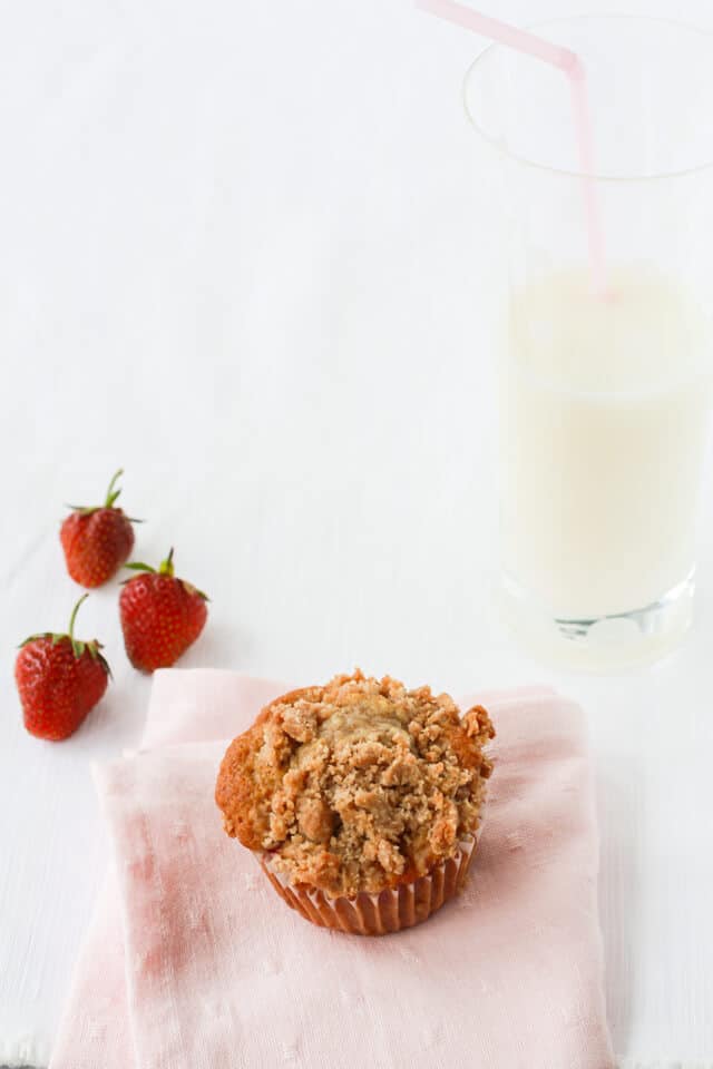 Strawberry rhubarb muffins with crumb topping and a glass of milk, pink napkin, 3 strawberries on the side