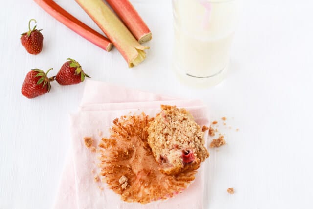 Half of a strawberry rhubarb muffins with crumble topping on a pink napkin with a glass of milk, and a few strawberries and stalks of rhubarb displayed nearby