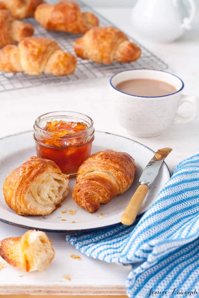 Croissant breakfast consisting of homemade croissants with a jar of orange marmalade served on a white enamelware plate with a blue rim, a knife, a blue and white striped napkin and a cup of coffee. Cooling rack of homemade croissants in the background