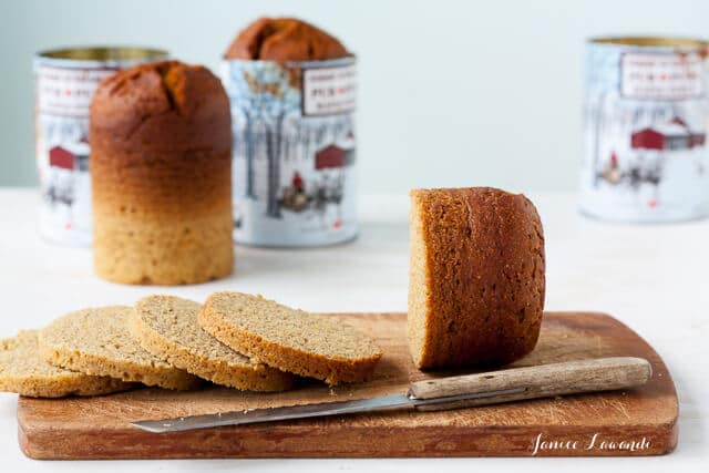 Sliced Quebec brown bread on a wood cutting board with serrated knife. Empty cans that were used to bake bread in the background