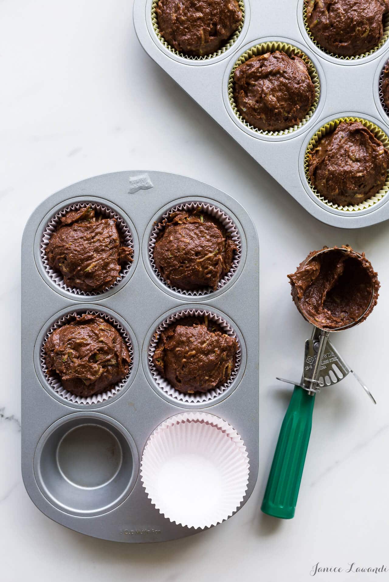 Lining a muffin pan with paper muffin liners and using a disher with a green handle to divide and scoop chocolate zucchini muffin batter