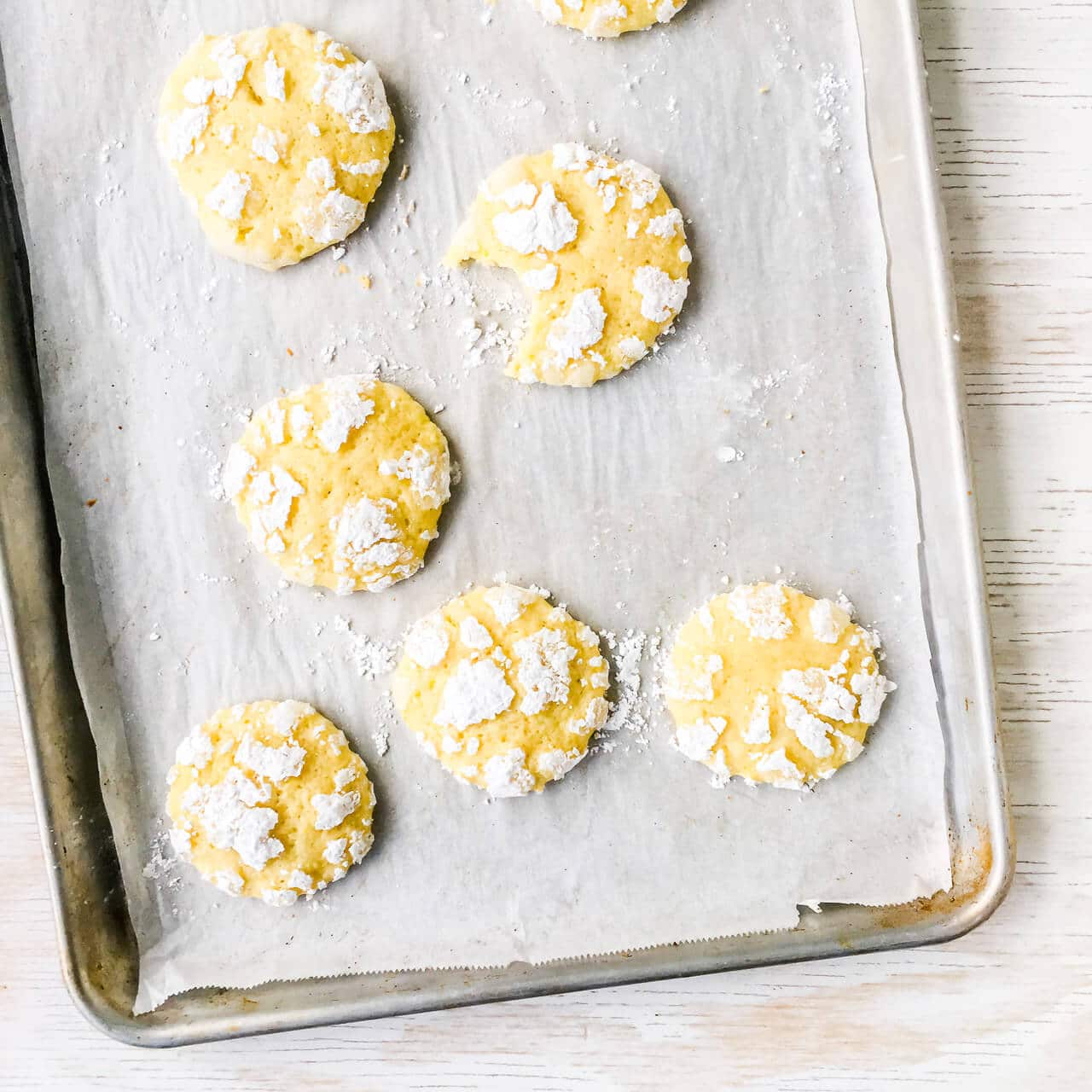 Lemon crackle cookies have a cracked texture of icing sugar on the surface of these yellow cookies, baked on a parchment-lined sheet pan.