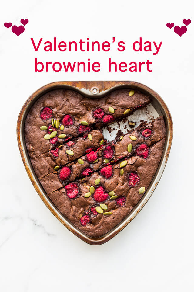 Heart-shaped dark chocolate brownie in a heart-shaped cake pan topped with raspberries and chopped nuts or pumpkin seeds