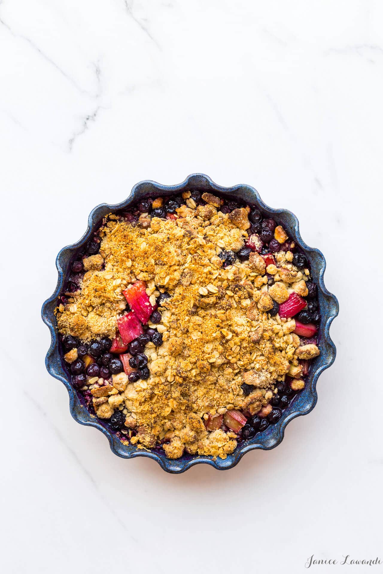 Blueberry rhubarb crumble with a marzipan oat crumble topping served in a round ceramic blue baking dish with a fluted edge
