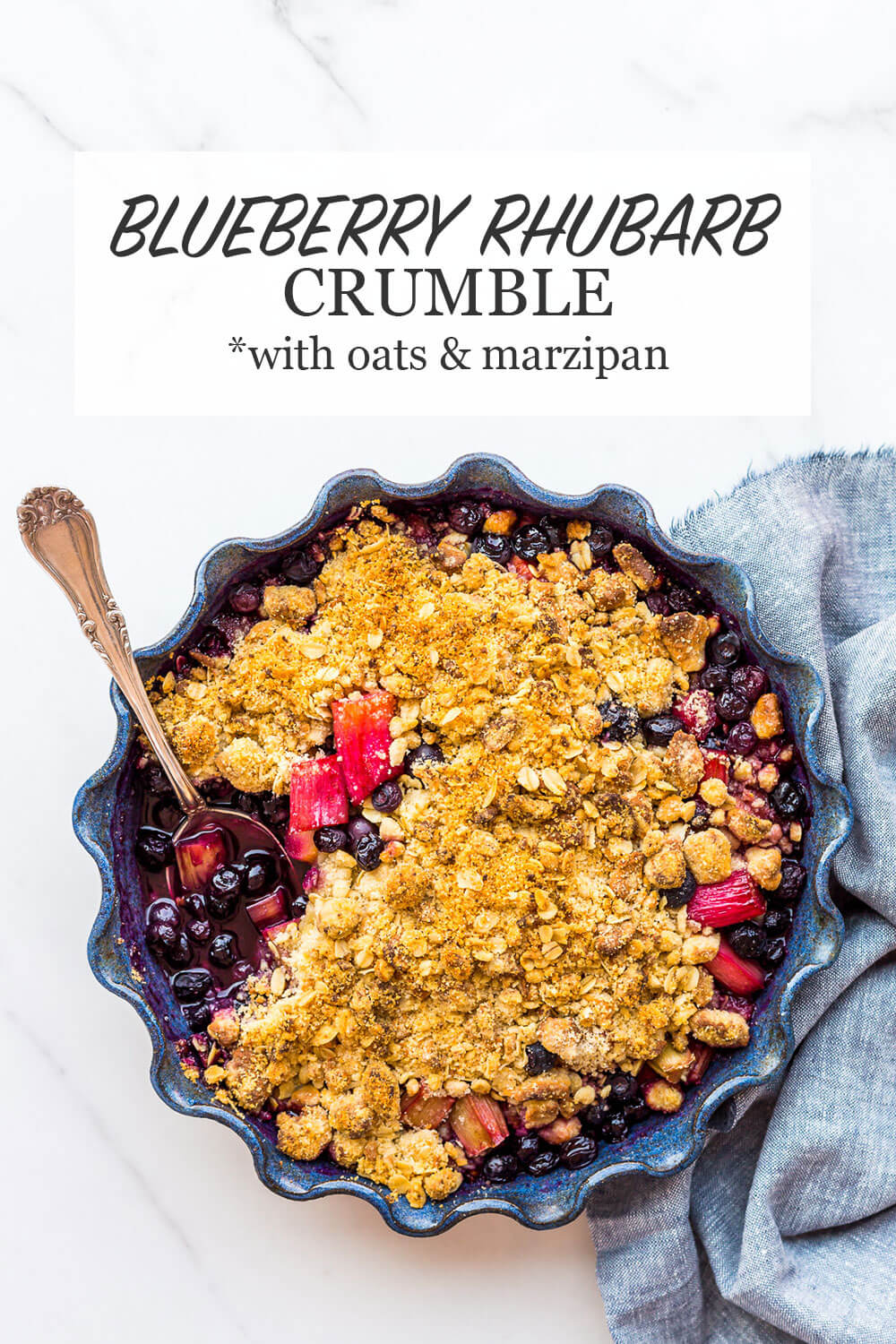 Blueberry rhubarb crisp with a marzipan and oat crumble topping baked in a blue ceramic baking dish with scalloped edges and served with a big serving spoon and blue linen 