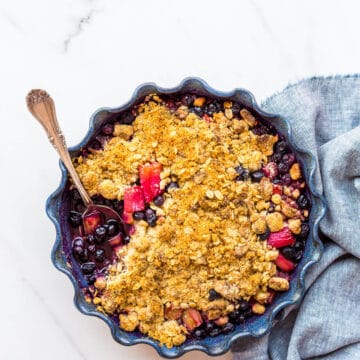 Blueberry rhubarb crumble with a marzipan oat crumble topping served in a round ceramic blue baking dish with a fluted edge. The blueberry rhubarb filling is very juicy.