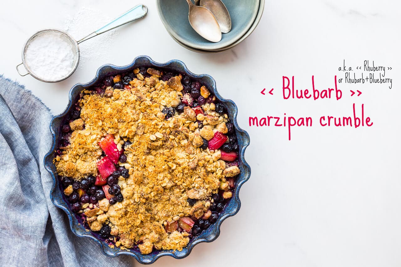 Blueberry rhubarb crumble with a marzipan oat crumble topping served in a round ceramic blue baking dish with a fluted edge