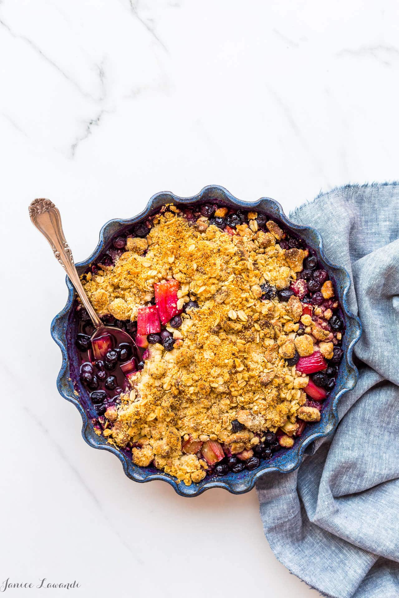 Blueberry rhubarb crumble with a marzipan oat crumble topping served in a round ceramic blue baking dish with a fluted edge. The blueberry rhubarb filling is very juicy.