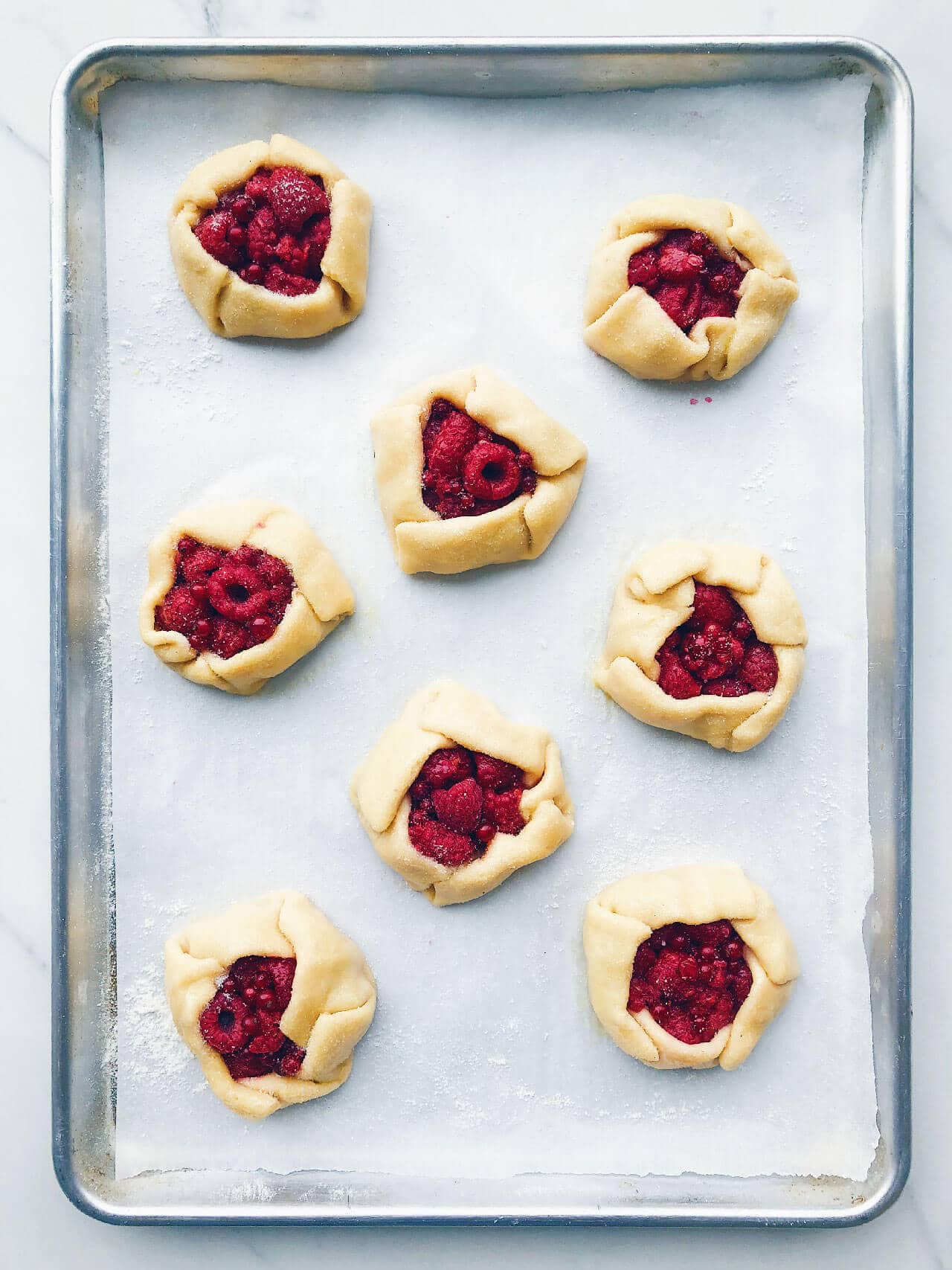 Homemade freeform mini raspberry galettes shaped and ready for the oven on a parchment-lined baking sheet