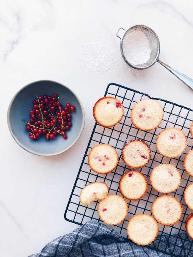 red currant financiers with a bowl of red currants and icing sugar in a small strainer for dusting over the little cakes on a black cooling rack