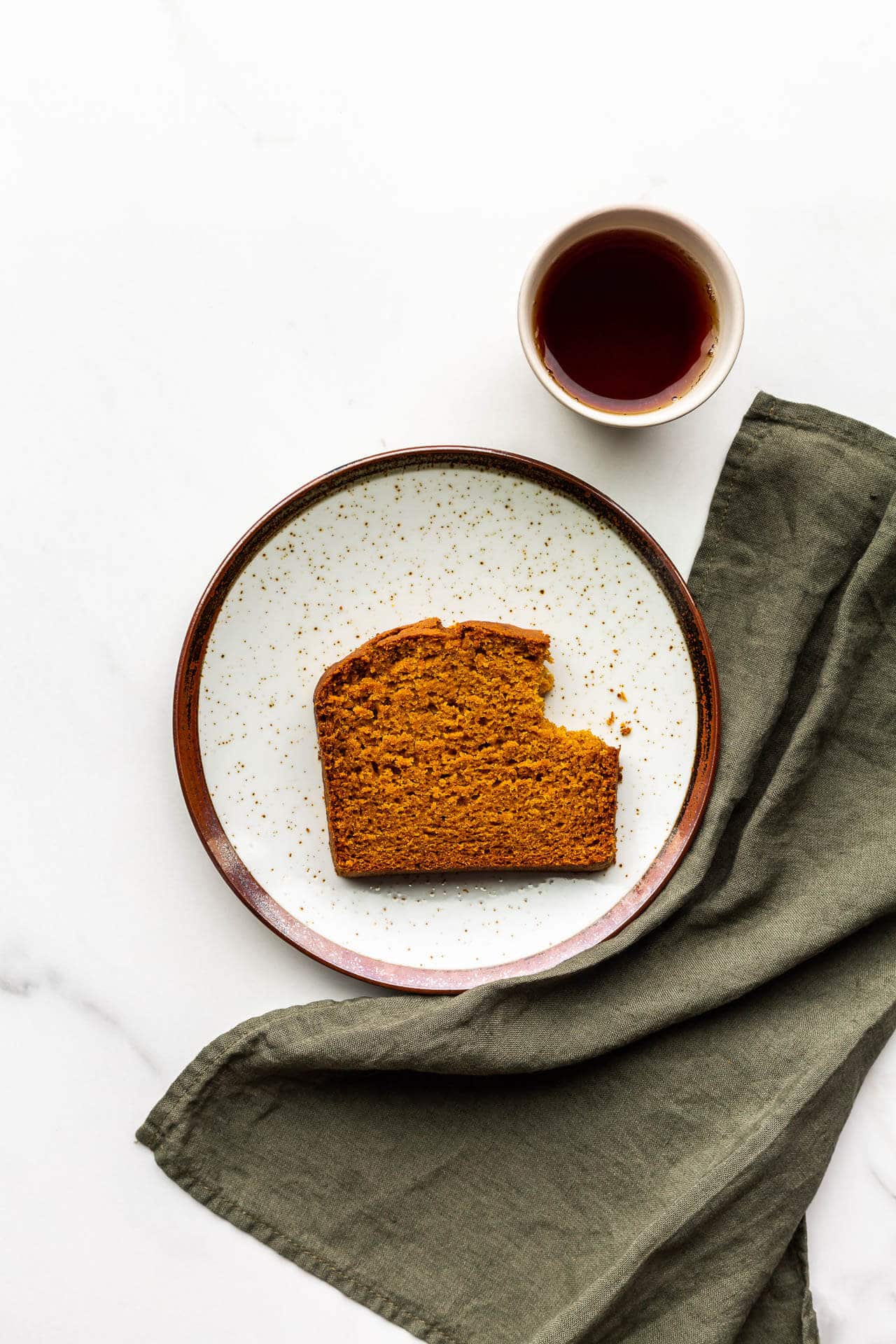 A slice of pumpkin spice bread with a bite taken out of it, with a green linen and a cup of tea on the side.