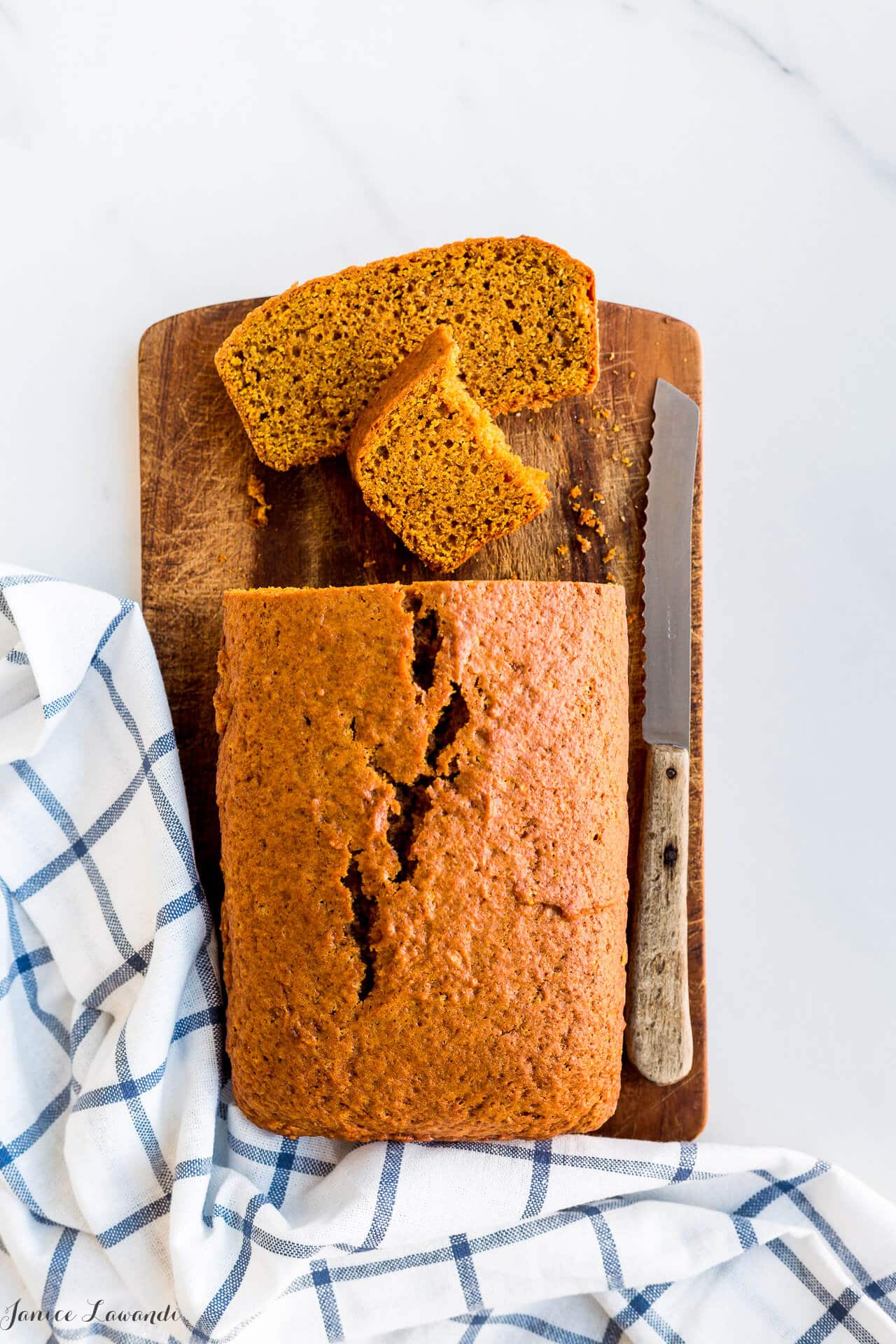 Slices of pumpkin loaf cake on a wooden cutting board with a serrated knife and a blue and white kitchen towel