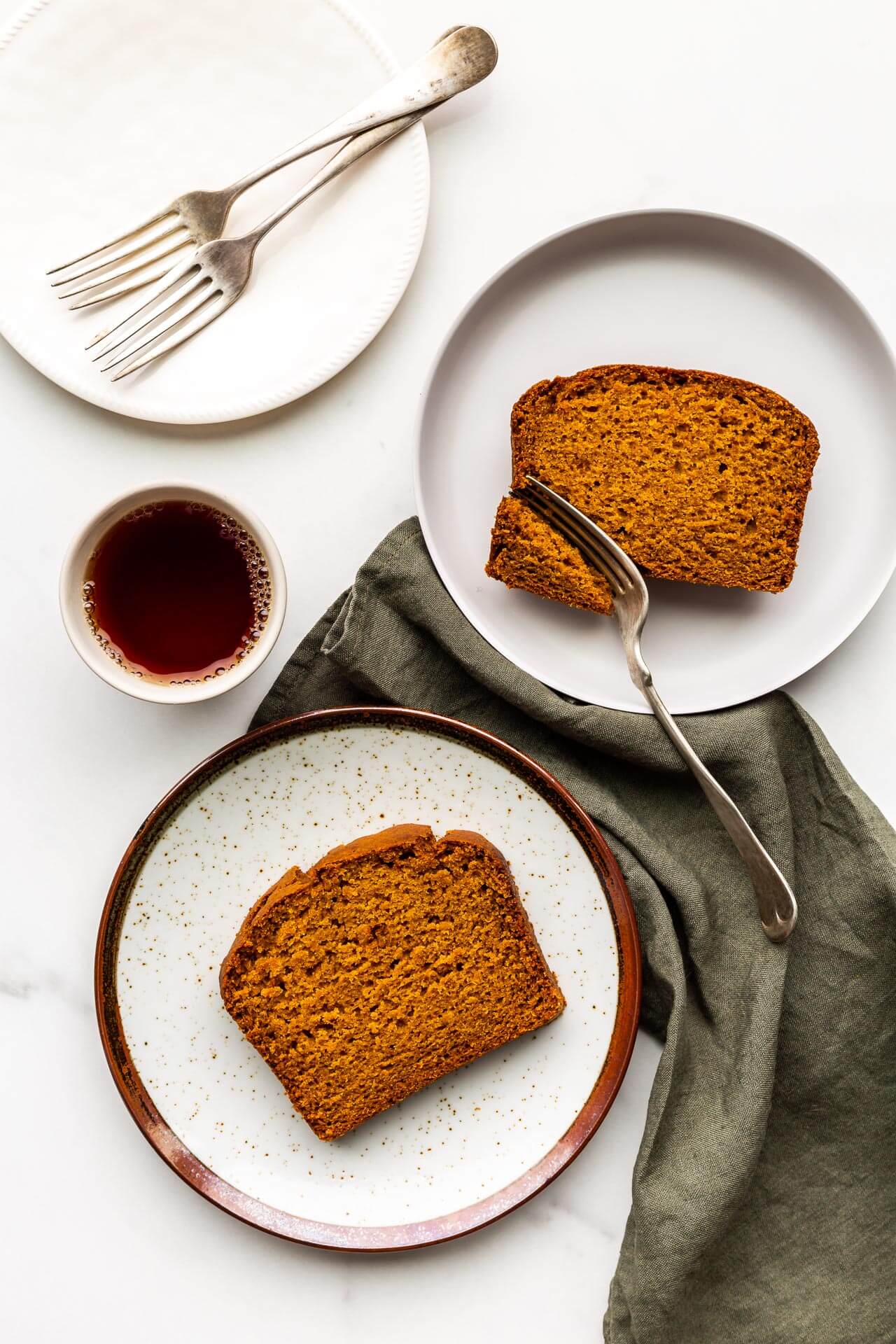 Slices of pumpkin bread served on speckled plates with a cup of tea and green linen