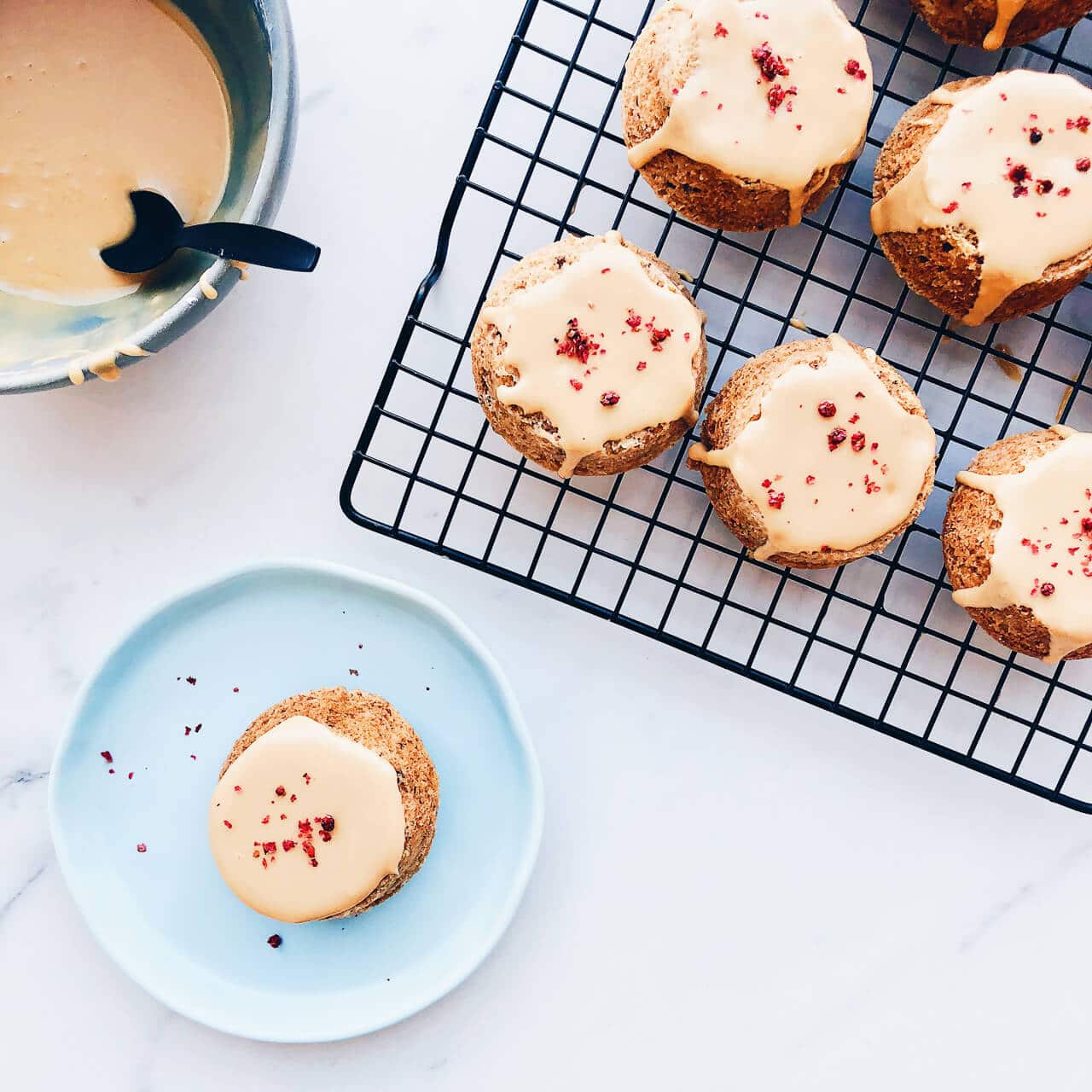 Little cardamom walnut cakes on a black cooling rack glazed with a coffee icing and topped with cracked pink peppercorns