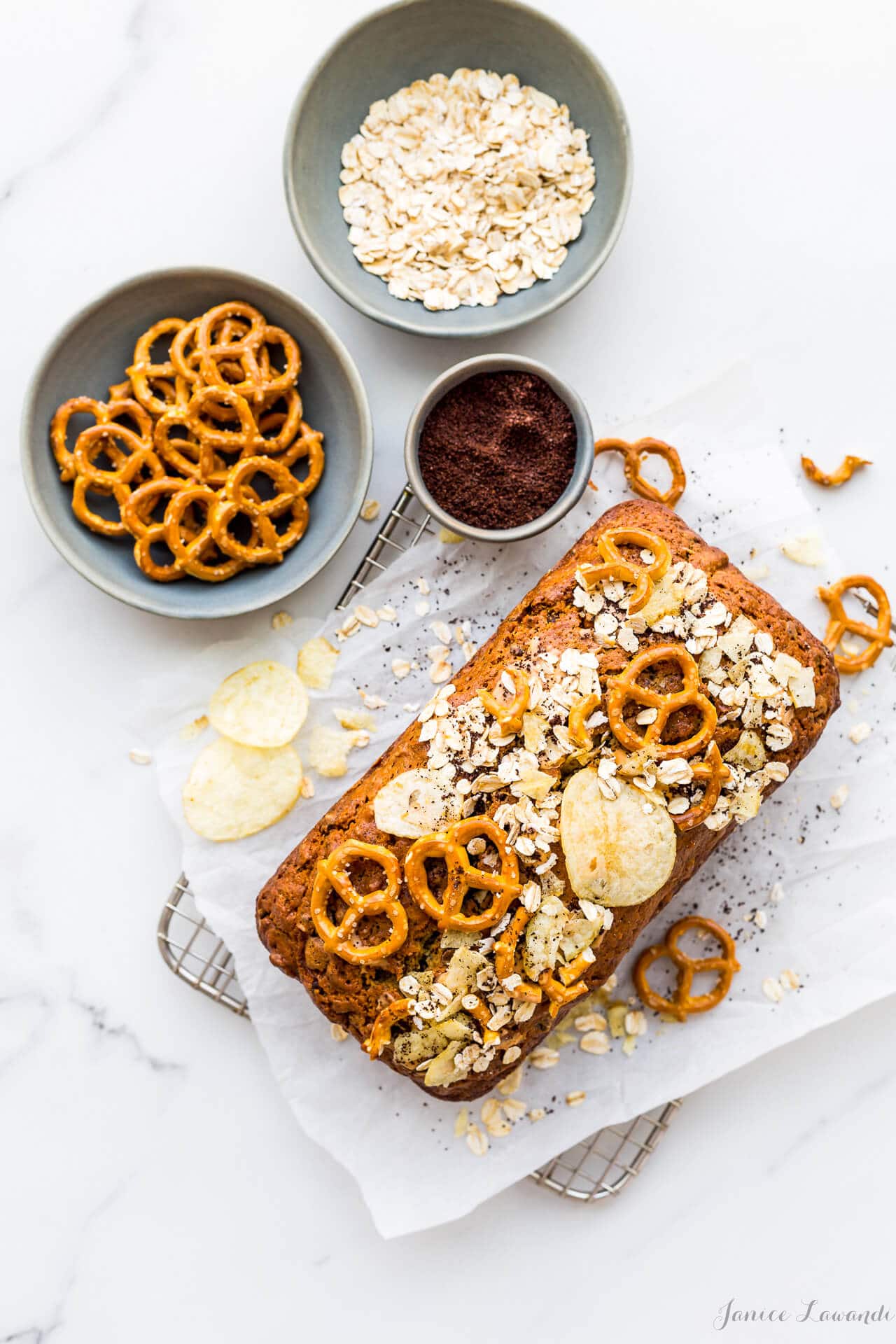 Compost pound cake on parchment on cooling rack topped with potato chips, oats, coffee grinds, and mini pretzels. With grey stoneware bowls filled with oats, pretzels, coffee grinds. 