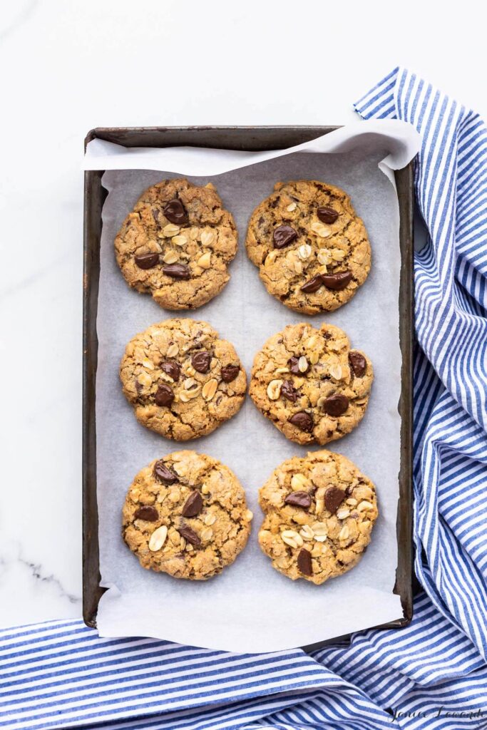 Freshly baked thick chewy oatmeal chocolate cookies with peanuts on a dark cookie sheet lined with parchment and a striped linen