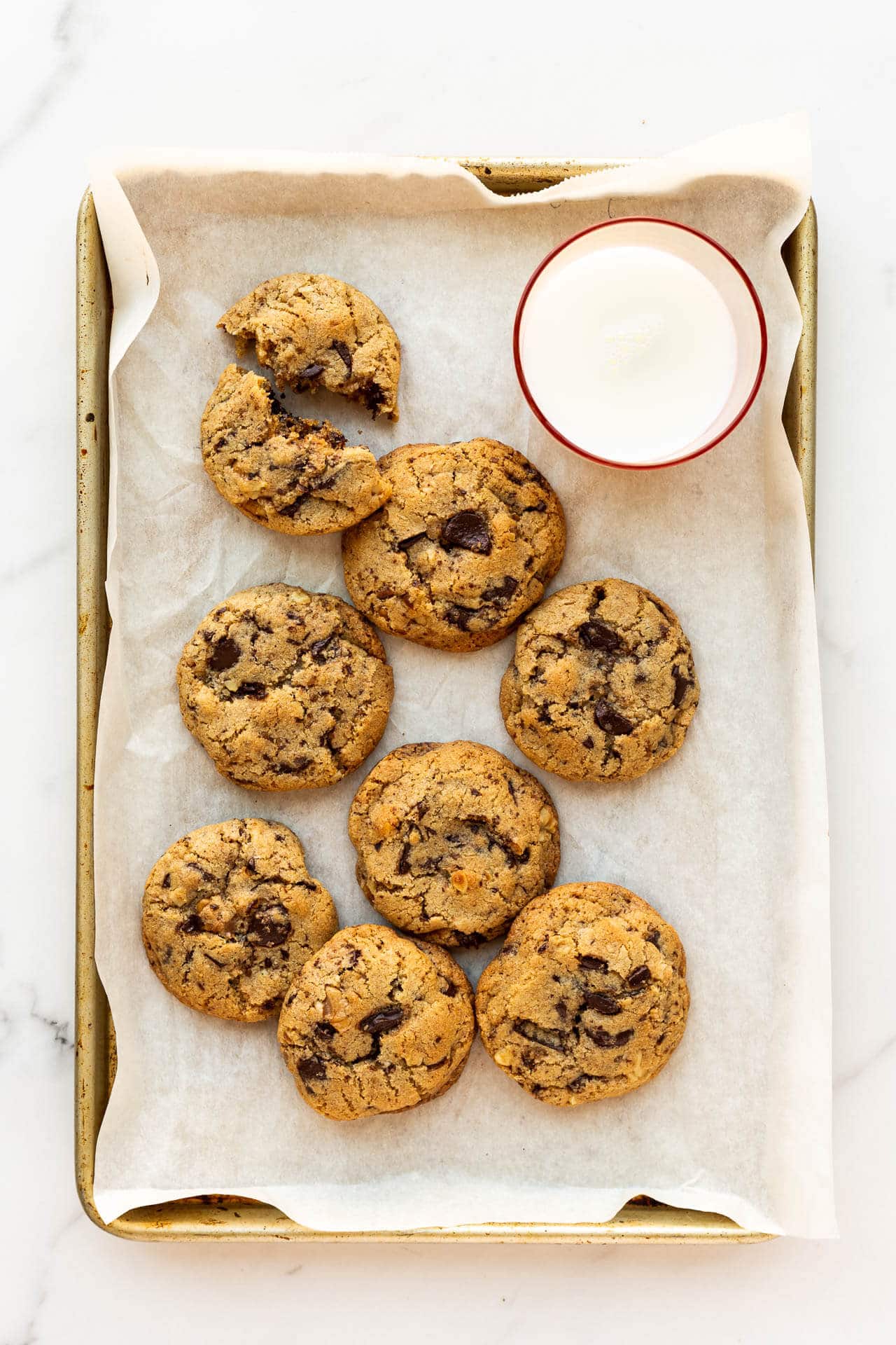 A parchment-lined tray of spelt flour chocolate chip cookies with a pink glass of milk.