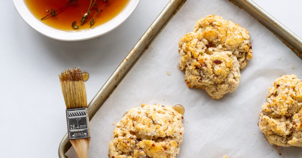 Golden baked biscuits on a sheet pan lined with parchment being glazed with a pastry brush and a small bowl of maple syrup with sprigs of thyme