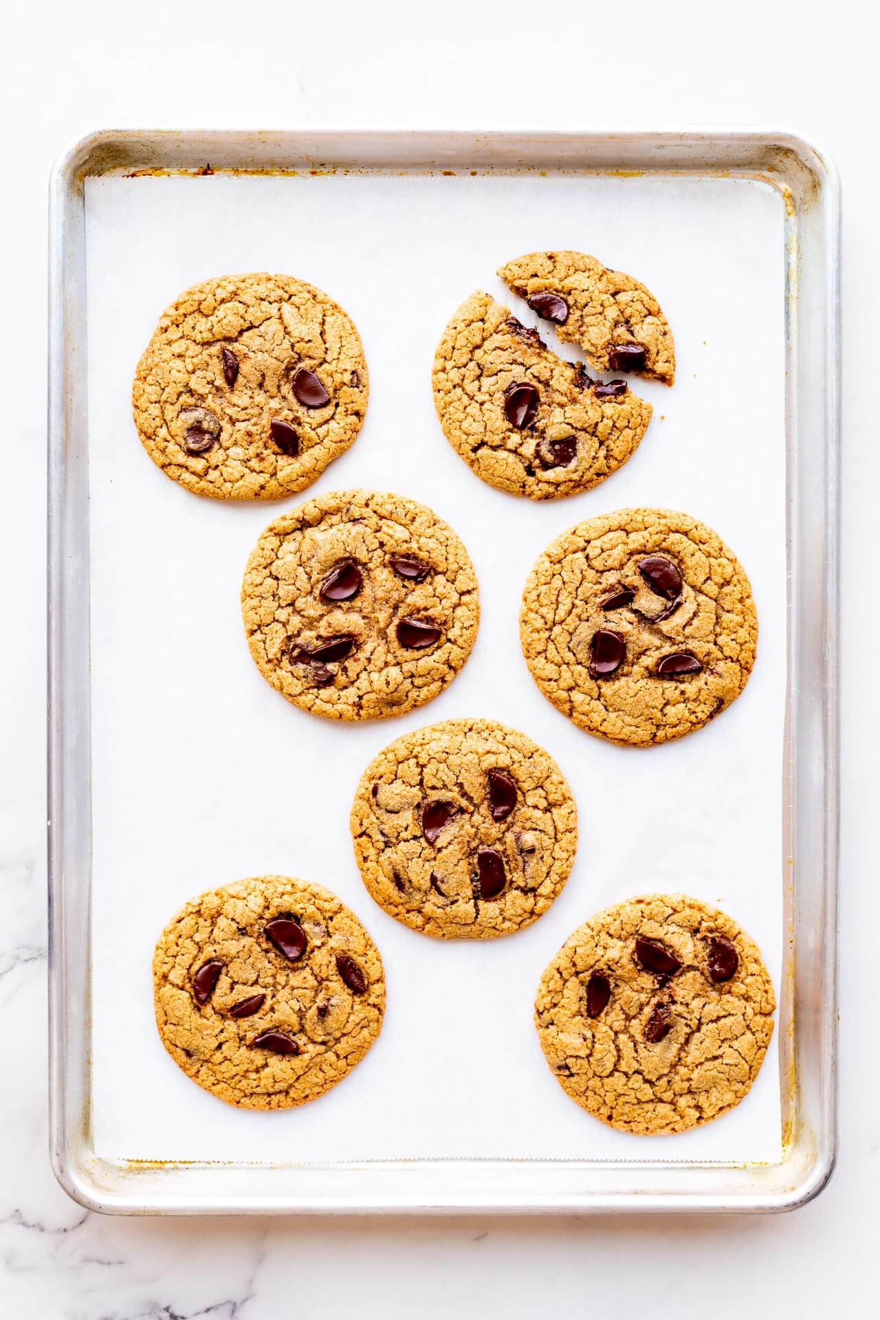 Freshly baked big chocolate chip cookies made with chunks of dark chocolate on a parchment-lined sheet pan with one cookie broken in two — 7 cookies on sheet pan