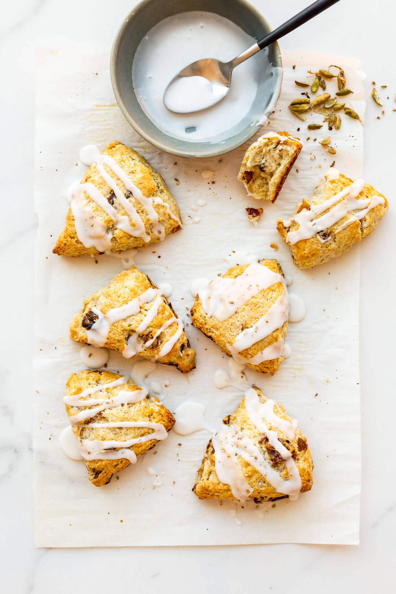 Six (6) glazed fruit scones with icing drizzled on from a bowl with a spoon, sprinkled with freshly ground cardamom from pods