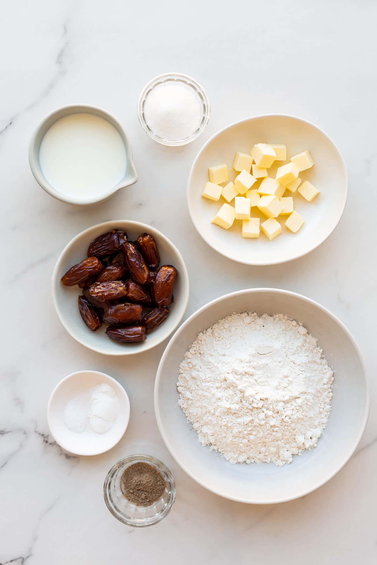 Ingredients to make date scones included cubed butter, salt, baking powder, cream, chopped pitted dates, sugar, flour, and ground cardamom all in bowls for mise-en-place