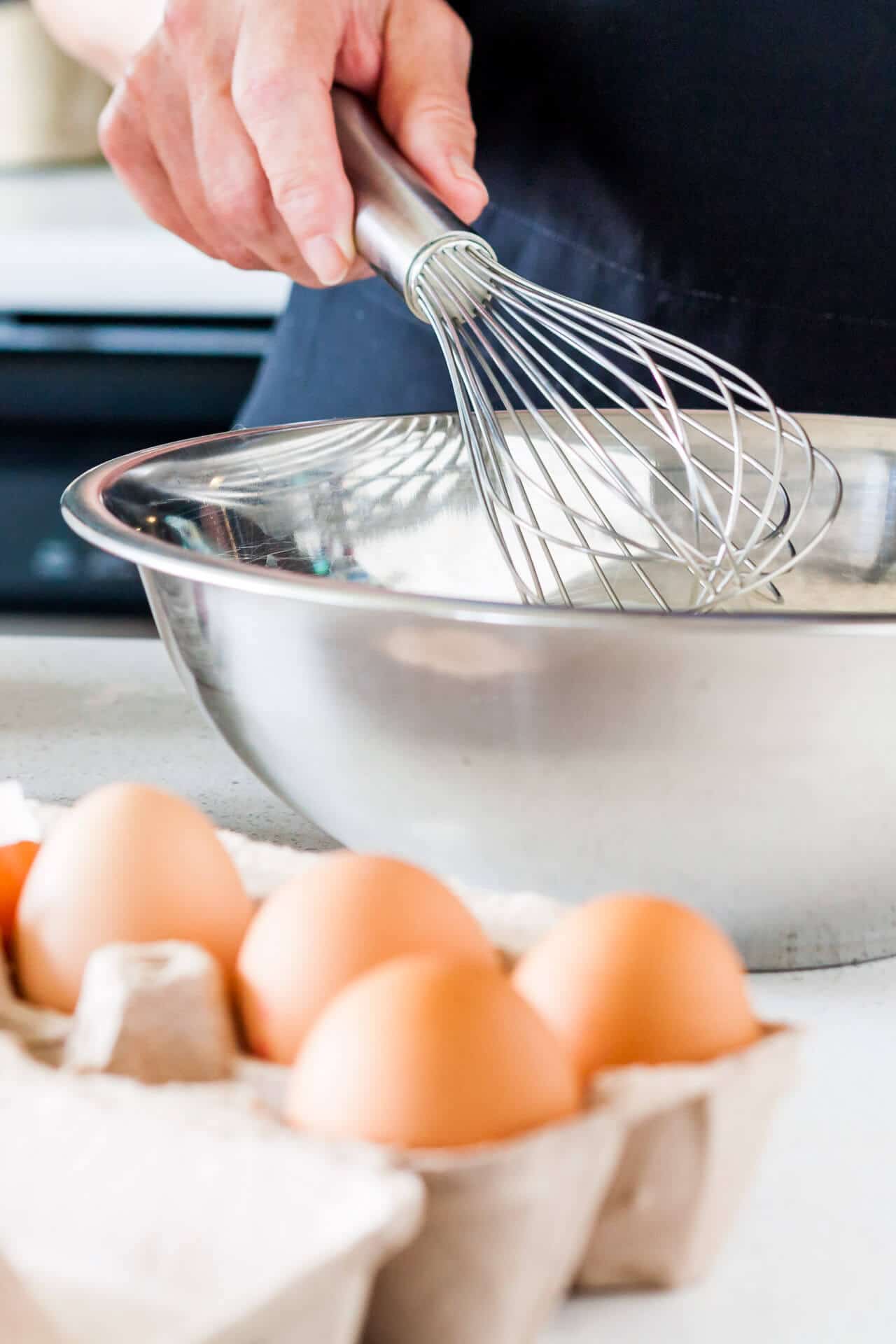 Whisking eggs by hand with a stainless steel whisk in a stainless steel bowl with a carton of brown eggs displayed next to the bowl