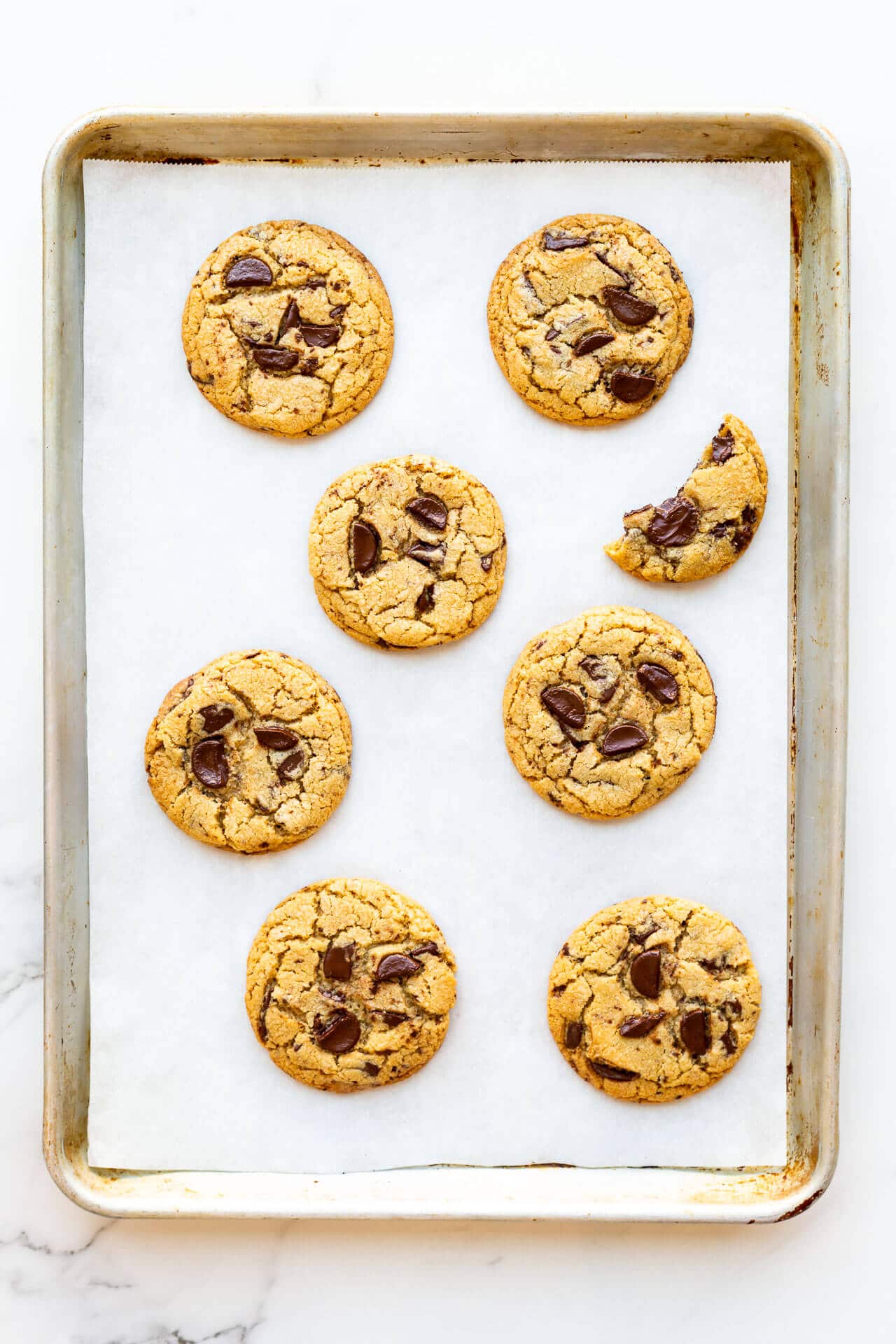 Thick chewy chocolate chip cookies baked with chunks of 70 % dark chocolate on a light sheet pan lined with parchment. One cookie is half-eaten