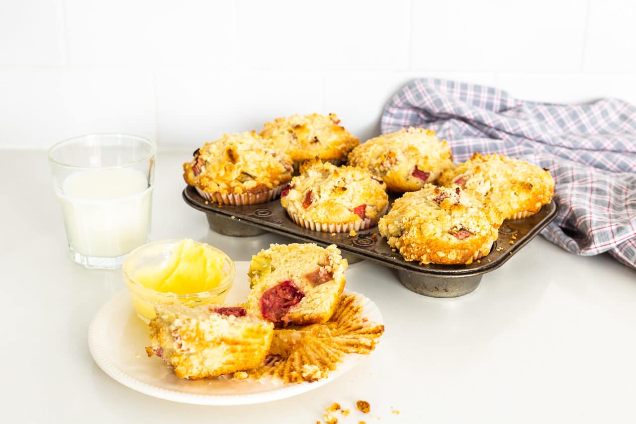 Vintage muffin pan with 6 bakery-style big pink rhubarb muffins with streusel on top, one muffin on a little white plate with a glass of milk and a serving of butter for the muffin