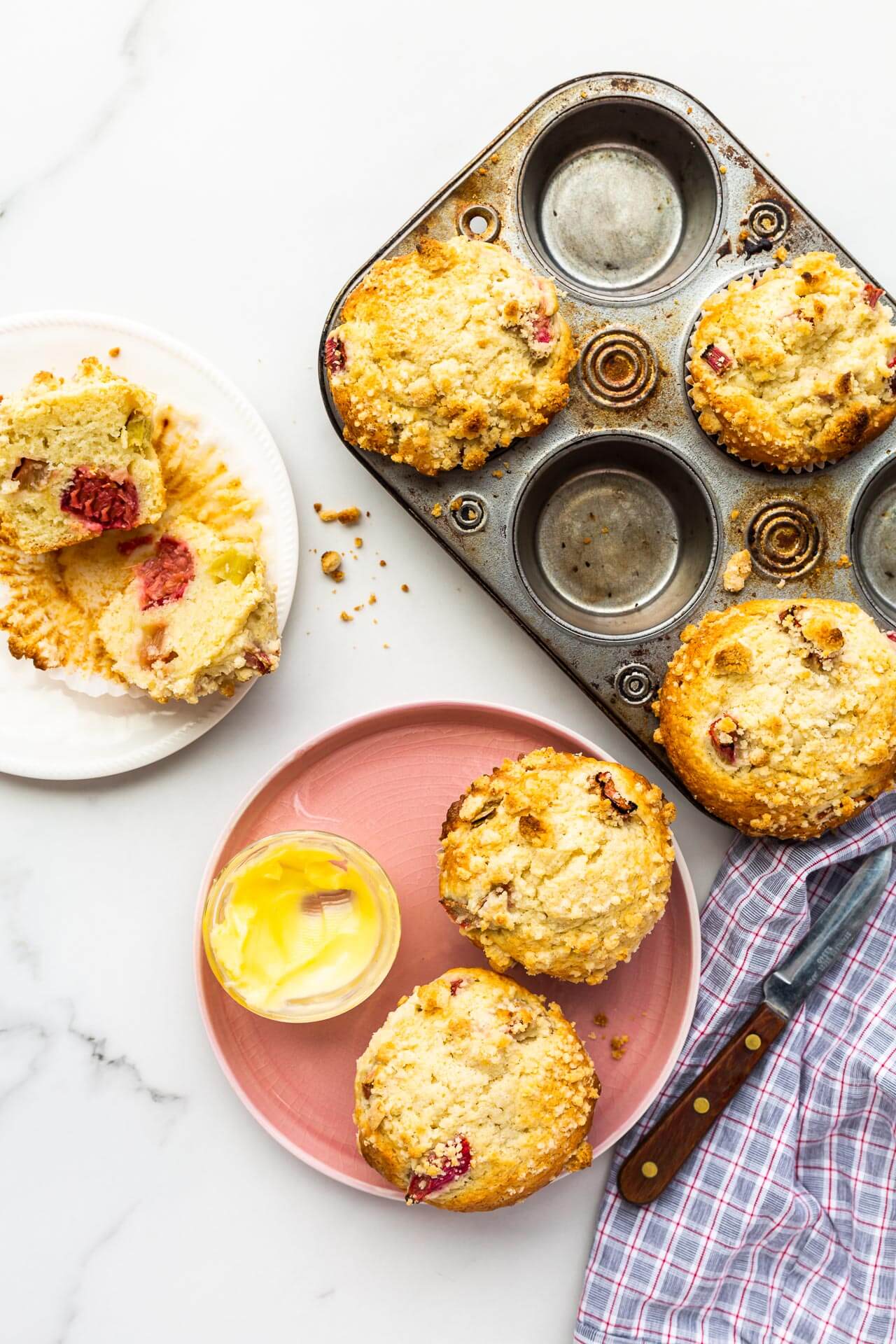 Vintage muffin pan half-full with rhubarb muffins with two on a pink plate and some butter, and another open on a little white plate