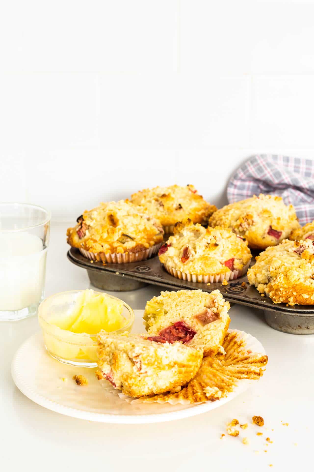 A vintage muffin pan of rhubarb muffins with one on a small plate, cut open to show pink rhurbarb inside, served with a glass of milk