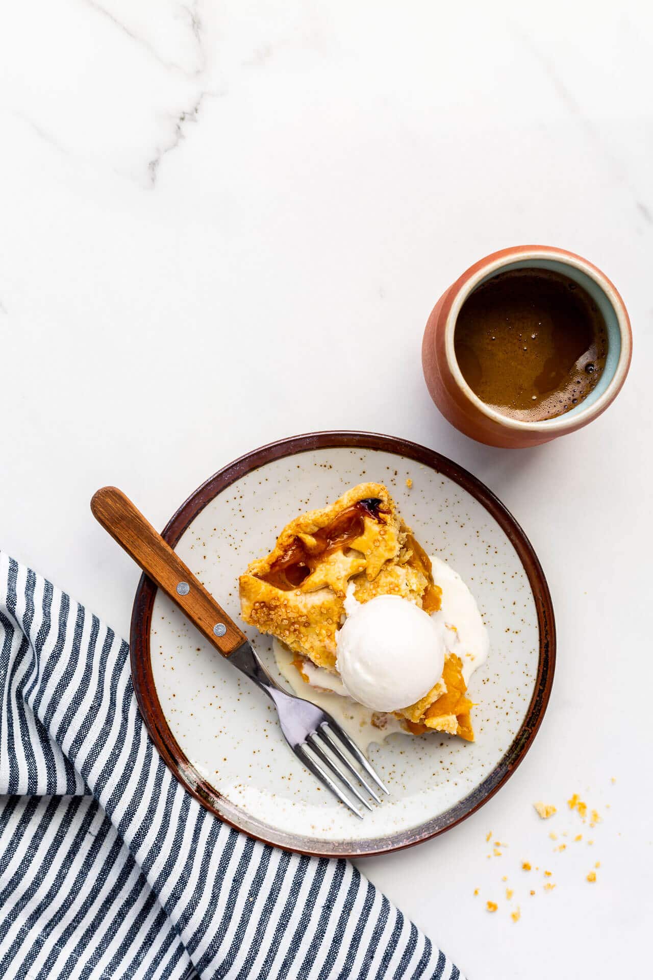 A slice of peach mango pie served on a ceramic plate with a cup of coffee and a blue and white striped linen