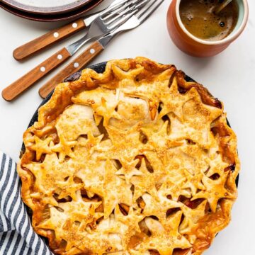 Cute pie with star design on top crust served with pile of plates and forks and a cup of coffee, striped linen