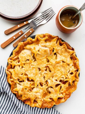 Cute pie with star design on top crust served with pile of plates and forks and a cup of coffee, striped linen
