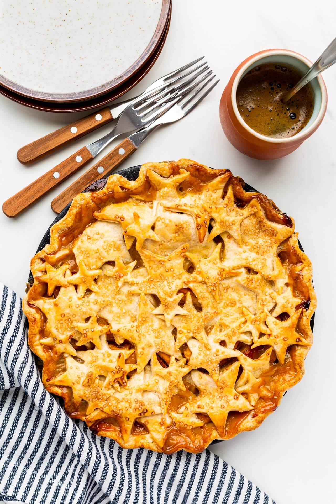 Cute pie with star design on top crust served with pile of plates and forks and a cup of coffee, striped linen