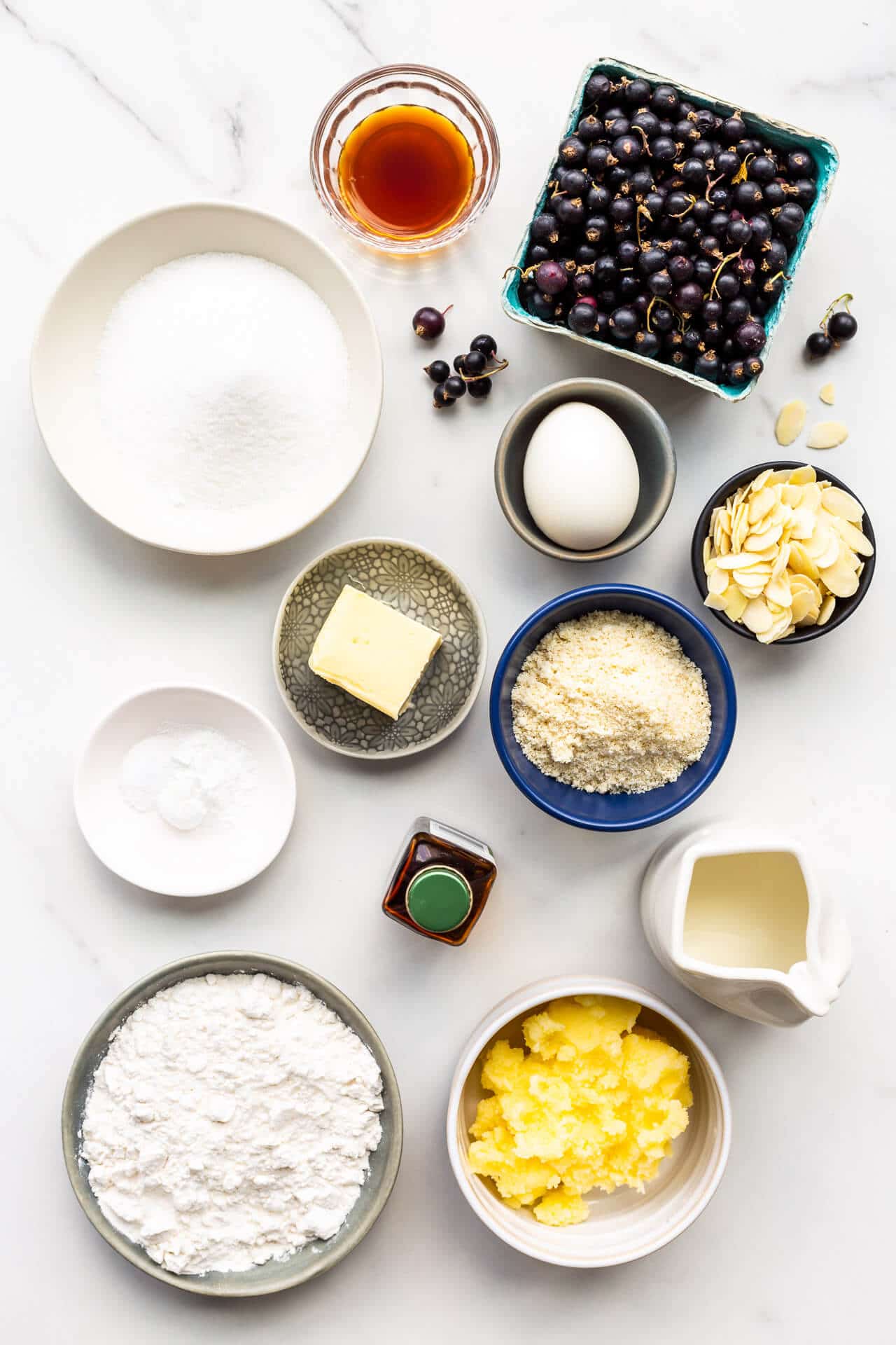 Ingredients for black currant cake measured out into small dishes, including black currants, vanilla extract, granulated sugar, egg, butter, ground almond, baking powder, salt, milk, flour, and a sugar paste to make a crispy topping