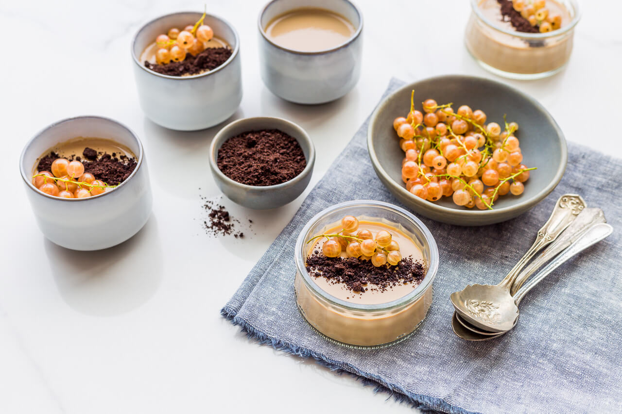 Cups of coffee panna cotta garnished with fresh white currants and Oreo cookie crumbs served on a blue chambray linen with spoons