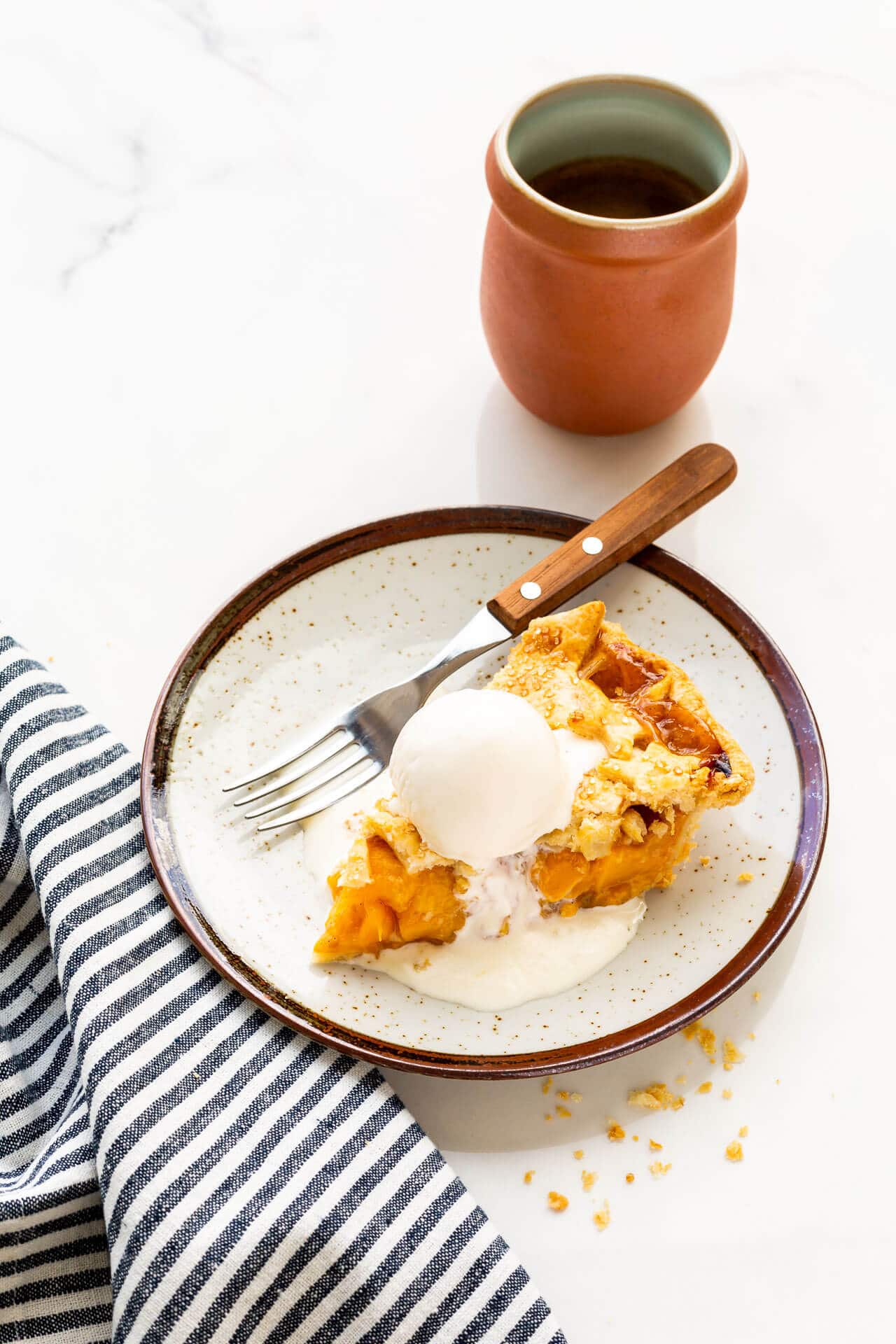 A slice of peach mango pie served à la mode with vanilla ice cream on a ceramic plate with a wood-handled fork and a cup of coffee, blue white striped linen
