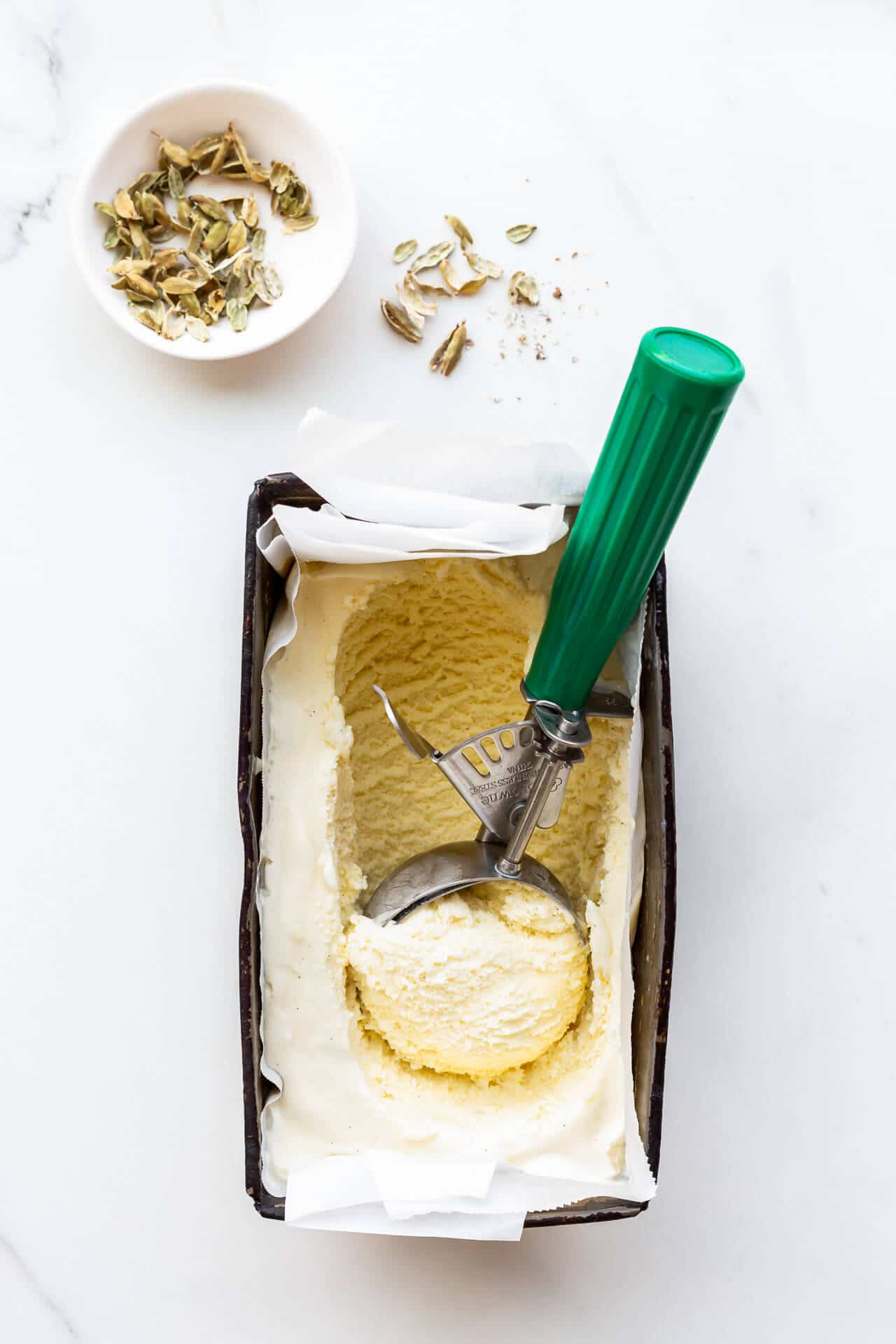 Scooping homemade cardamom ice cream with a disher. Small bowl of cardamom pods nearby