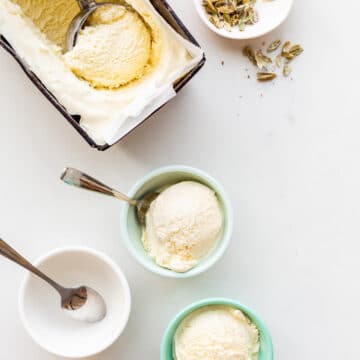 Two bowls of cardamom ice cream with spoons ready to eat with the pan of cardamom ice cream and a scoop ready to fill the third bowl. Little bowl of cardamom pods on the side.