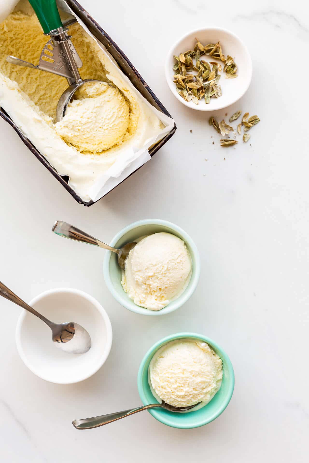 Two bowls of cardamom ice cream with spoons ready to eat with the pan of cardamom ice cream and a scoop ready to fill the third bowl. Little bowl of cardamom pods on the side.