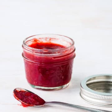 A jar of homemade strawberry red currant jam with a spoonful on the side and the metal band