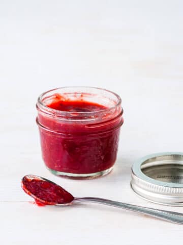 A jar of homemade strawberry red currant jam with a spoonful on the side and the metal band