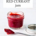 A jar of red currant jam made with strawberries with spoon of jam on side and metal bands