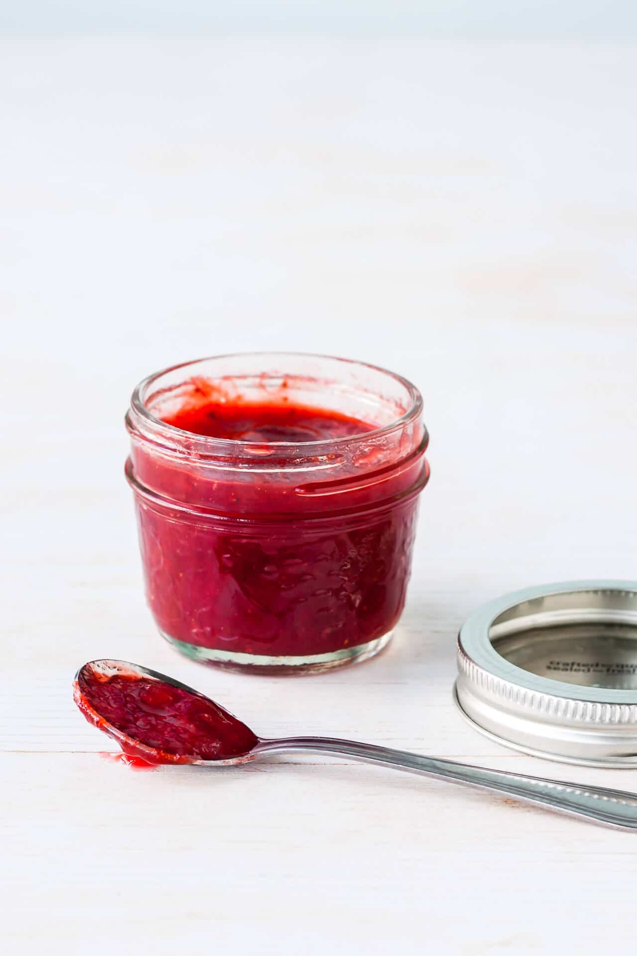 A jar of homemade strawberry red currant jam with a spoonful on the side and the metal band