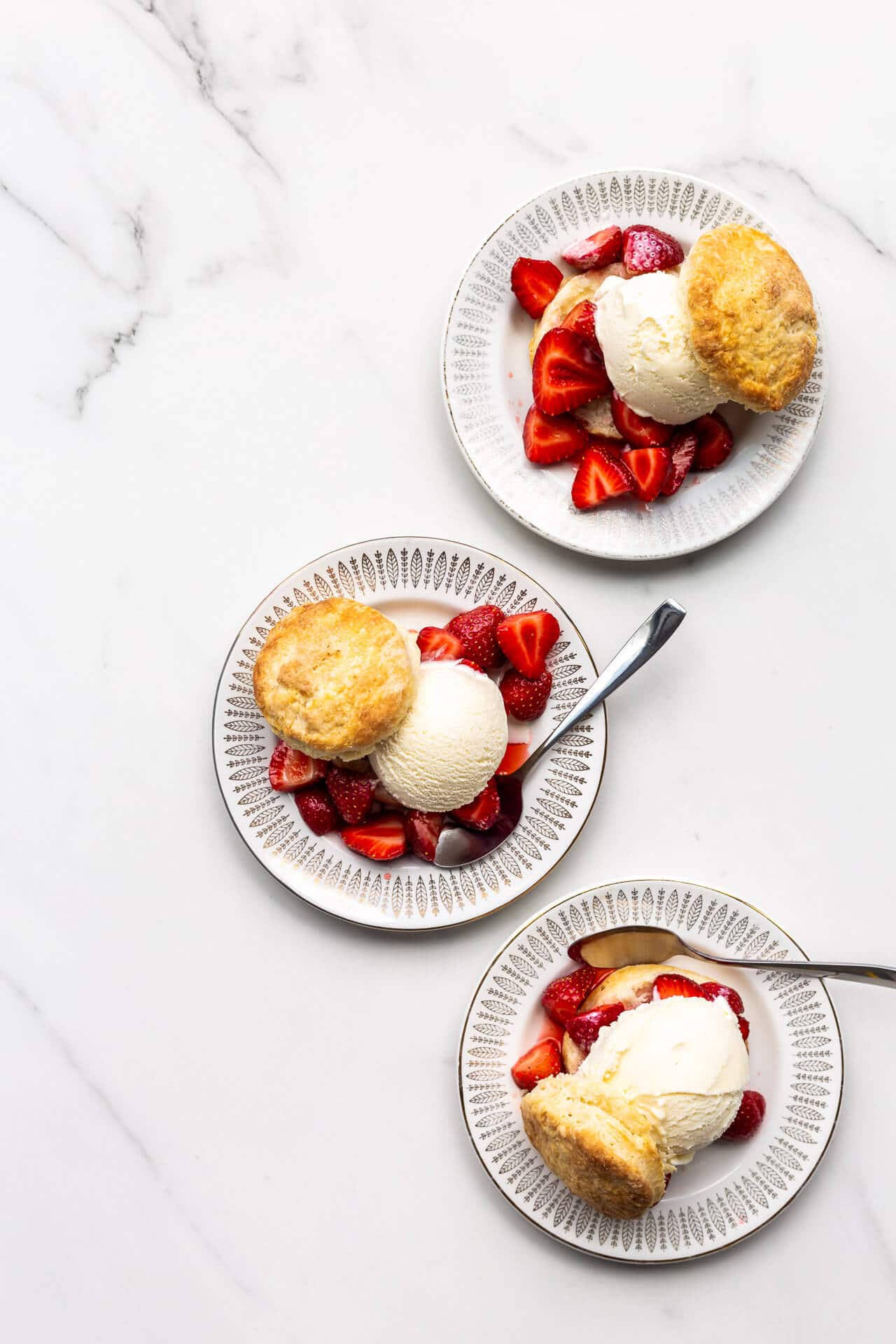 Three ice cream strawberry shortcakes on small dessert plates on white marble surface